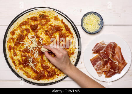 Frau Hand ist das Streuen Pizza mit Käse. Rezept Schritt für Schritt Pizza Capricciosa flatlay auf weißem Holz Stockfoto