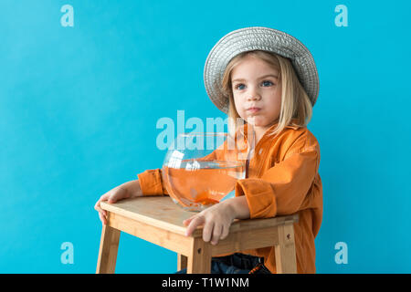 Kid in Silber Hut und orangefarbenen T-Shirt holding Aquarium mit Goldfischen und Wegsehen auf Blau isoliert Stockfoto