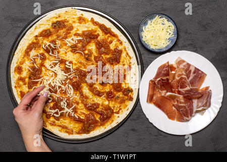 Frau Hand ist das Streuen Pizza mit Käse. Rezept Schritt für Schritt Pizza Capricciosa flatlay am grauen Stein Stockfoto