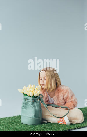 Cute kid mit gekreuzten Beinen riechenden weißen Tulpen im Beutel isoliert auf Grau Stockfoto