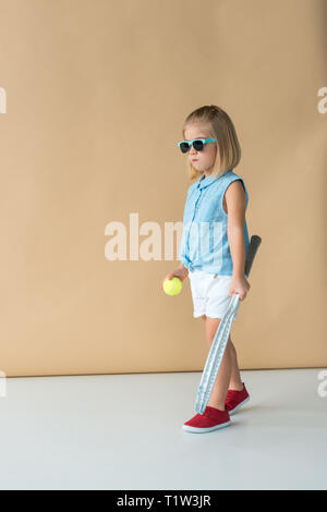 Niedliche Kind in der Sonnenbrille, T-Shirt und Shorts mit Schläger und Ball auf beigen Hintergrund Stockfoto