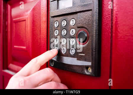 Hand drückt eine Taste Klingel- oder Sprechanlage auf rote Tür. Sicherheitskonzept. Stockfoto