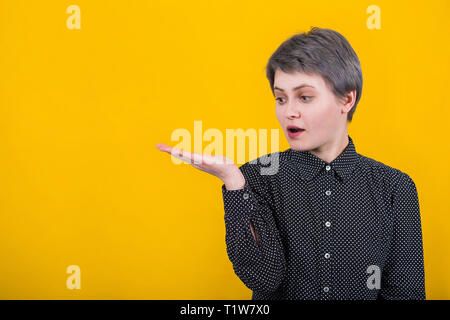 Gut aussehenden jungen kaukasischen Frau legere schwarzes T-Shirt, hält die Hand erhoben wie das Halten einer imaginären Objekt, Schauen, Staunen, isoliert über y Stockfoto