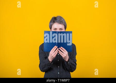 Glückliche Frau Buch Leser versteckt sich hinter einem blauen Lehrbuch, hat freudige Ausdruck und positive Sicht, findet heraus neue, interessante Hobby. Weibliche hipster Studi Stockfoto