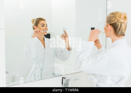 Selektiver Fokus der schönen Frau in weißen Bademantel Zähneputzen und die selfie im Badezimmer Stockfoto