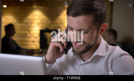 Closeup Schießen von Erwachsenen kaukasischen Geschäftsmann Tippen auf dem Laptop und dem ungezwungenen Gespräch am Telefon freundlich lächelnd drinnen im Büro Stockfoto