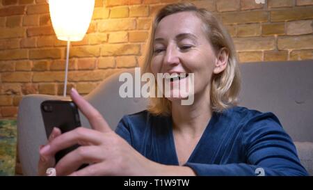 Close-up Portrait von freudiger Senior kaukasischen Dame aufpassen in Smartphone und lachend in gemütlicher Atmosphäre. Stockfoto