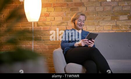 Portrait von Senior kaukasischen Dame sitzt auf einem Sofa und Beobachten in Tablet gerne in gemütlicher Atmosphäre. Stockfoto