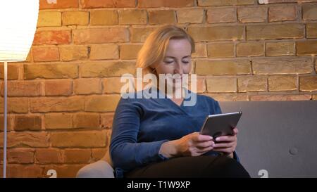 Portrait von Senior kaukasische Frau sitzt auf einem Sofa und Beobachten in Tablet aufmerksam in gemütlicher Atmosphäre. Stockfoto