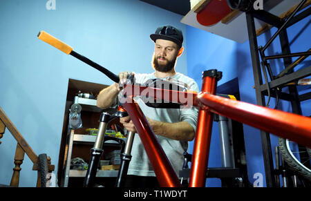 Ein Mann Fahrradmechaniker baut ein Mountainbike in seiner Werkstatt. Ein Mann mit einer Kappe mit einer Bart. Der Mechaniker rollt das Rad mit dem Fahrrad Rahmen Stockfoto