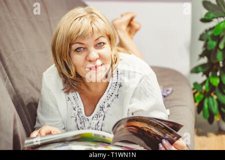 Eine ältere Frau liest eine Zeitschrift auf der Couch zu Hause liegen Stockfoto