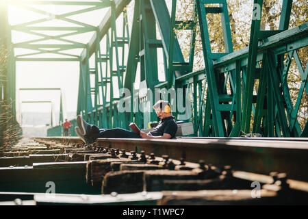 Junge lächelnde Bauarbeiter Mann sitzt mit einem Buch auf einer alten Eisenbahnbrücke aus Stahl Stockfoto