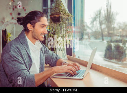 Glücklich lächelnde Mann am Laptop mit der Eingabe. Portrait von ein hübscher Kerl trägt formalen weißes Hemd und grauen Bluse sitzt in der Nähe der Fenster am Tisch im Wohnzimmer oder Stockfoto