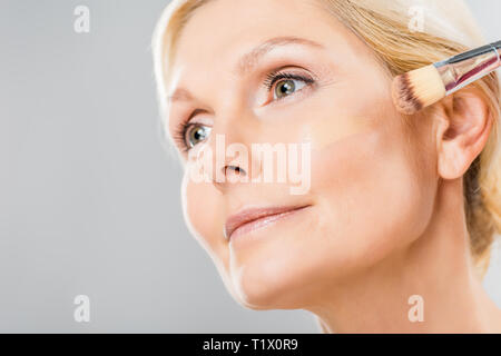 Schöne und reife Frau weg schauen und Anwendung von Make-up-Grundlage mit Kosmetik Pinsel isoliert auf Grau Stockfoto