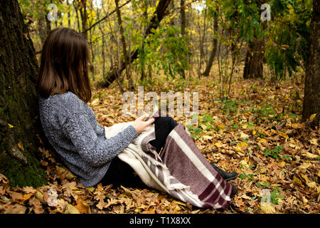 Junge schöne Mädchen in einer grauen Jacke in den Wald in der Nähe einen grossen Baum mit einem Mobiltelefon. Stockfoto