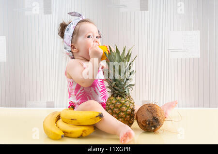 Süße kleine Mädchen in rosa Shirt sitzen unter tropischen Früchten auf den Tisch und das Essen einer Zitrone, gesehen von der Seite Stockfoto