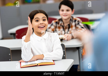 Aufgeregt Schulmädchen mit Buch Hand heben während der Lektion im Klassenzimmer Stockfoto