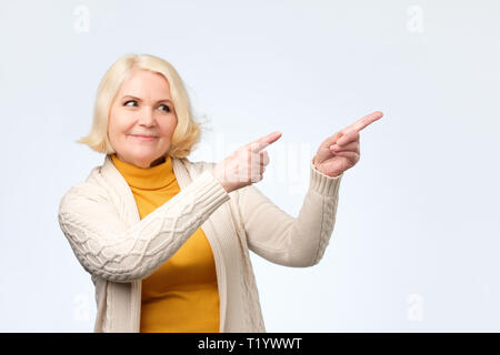 Gerne blonde ältere Frau lächelnd Pointing Finger weg, die etwas Interessantes und Spannendes in Studio Wand Stockfoto