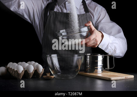 Man Zucker in die Schüssel auf schwarzen Hintergrund ausgießen. pie Konzept zu machen. Stockfoto