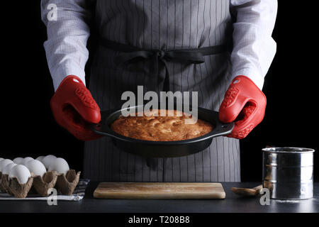Mann in Schürze Holding frisch gebackene Torte auf dunklem Hintergrund. Rezept od-Torte oder Kuchen. Stockfoto