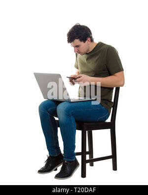 In voller Länge Porträt des jungen Mannes Freiberufler auf Stuhl mit seinem Laptop und Mobiltelefon in einem Zeit sitzen, wodurch verschiedene Berechnungen und Vergleichen Stockfoto