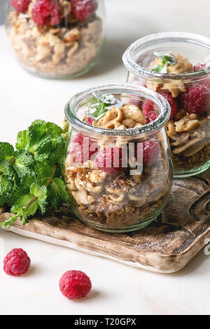 2-in-1-Dessert in Töpfen. Keks, Kaffeesahne, Muttern, Himbeeren, frischer Minze. Auf Keramik Tablett mit Beeren und Grüns über weißem Marmor Tisch serviert. Stockfoto