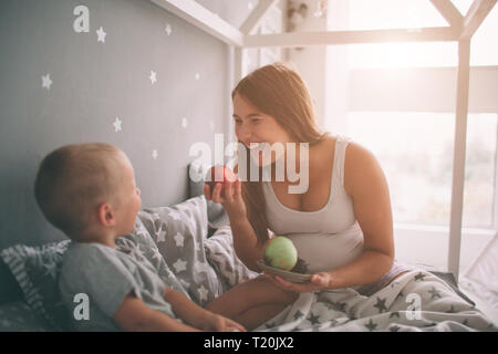 Schwangere Mutter und kleinen jungen Sohn isst einen Apfel und Pfirsich im Bett t Start in den Morgen. Casual Lifestyle im Schlafzimmer Stockfoto