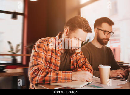 Ein Bild von bärtigen Kerl schreiben Sie Notizen in sein Notizbuch, während sein Freund ist das Lesen eines Artikels auf dem Telefon. Sie sind sehr beschäftigt und konzentrierten Stockfoto
