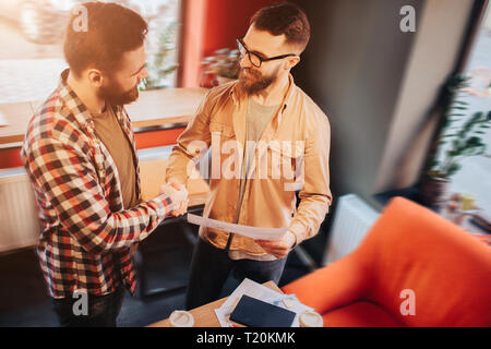 Zwei Hipster, legere Kleidung tragen in einem slall Cafe und schütteln ihre Hände. Einer von ihnen ist holdnig ein Dokument in der Hand. Beide sind Stockfoto