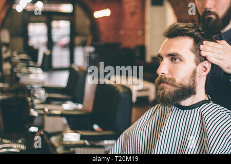 Bärtiger Kerl sitzt im Salon. Seine hairstylist ist Modellierung sein Haar. Kerl sucht gerade nach vorn Stockfoto