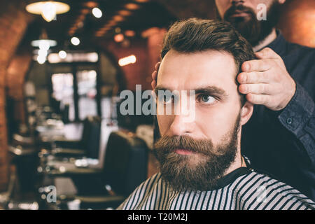 In der Nähe von schweren und bärtiger Mann im Sessel sitzen und gerade nach vorne zu schauen. Seine Friseur ist Modellierung sein Haar Stockfoto