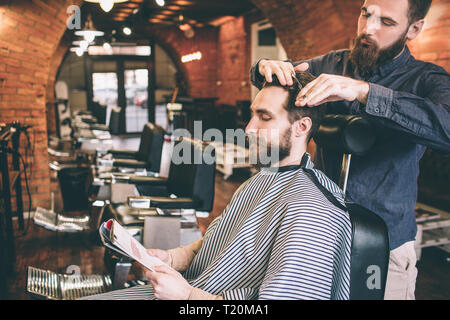 Der Kunde sitzt im Sessel und lesen Magazin während der Friseur schneiden die Haare. Er tut dies sehr professionell Stockfoto