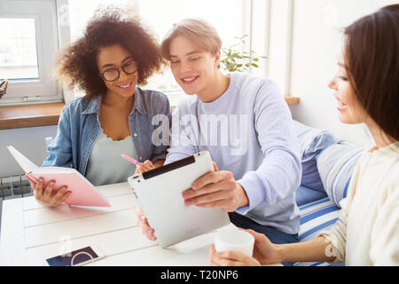 Schönes Bild von Guy zeigen etwas auf Tablet für ein Mädchen im blauen Hemd. Ihr Freund im weißen Pullover sitzt neben Ihnen und Halten einer Tasse te Stockfoto