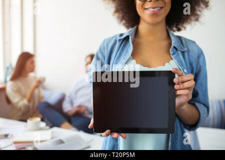 Schnittansicht der afro-amerikanische Mädchen steht und Demonstration eines schwarzen Tablette. Es sind Ihre Freunde am Tisch sitzen und miteinander reden Stockfoto