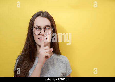 Innen- schuss von glücklichen Frau in der Brille sieht aus Kamera, hat faszinierende Blick, fragt ruhig zu sein, in T-Shirt gekleidet, steht vor gelbem Hintergrund Stockfoto