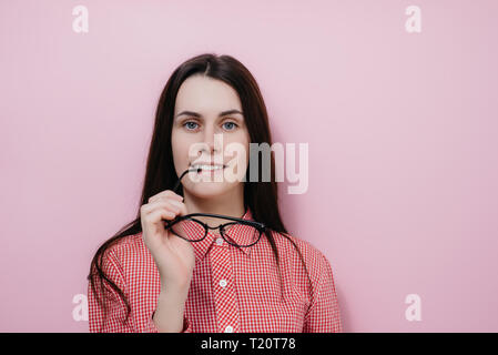 Porträt der glückliche junge Frau hält die Brille in der Nähe von Lippen, freut sich recieving Kompliment, legere Kleidung, über rosa Hintergrund isoliert Stockfoto