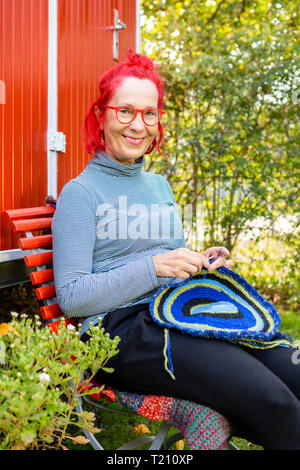 Portrait von lächelnden älteren Frau mit rot gefärbten Haaren vor der Roten Anhänger Sitzen im Garten Häkeln Stockfoto