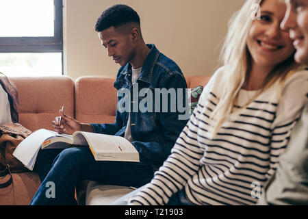 Afrikanische männliche Kursteilnehmer Notizen beim Lesen von Büchern im Sofa sitzen am College Campus. Studenten studieren auf dem Campus Gebäude mit Mädchen sitzen in her Stockfoto