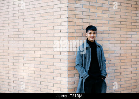 Portrait von lächelnden jungen Mann mit grauen Mantel und schwarzen Rollkragenpullover Pullover gegen die Wand lehnt Stockfoto