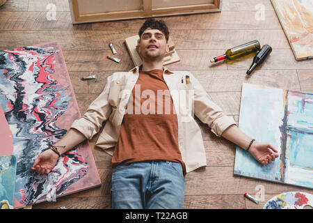 Erschöpft Künstler liegen auf dem Boden in der Malerei studio, von Gemälden, leere Flaschen und zeichnen Utensilien umgeben Stockfoto