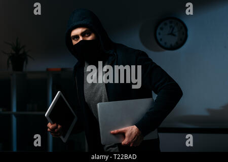 Dieb in Maske stehlen Laptop und einer Tablette und Wegsehen Stockfoto