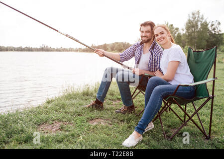 Ein Bild der ehrfürchtige junge Leute zusammen am Fluss Ufer sitzen und lächelnd vor der Kamera. Guy ist riesig und lange Fische - Rod, während seine Frau ist leani Stockfoto
