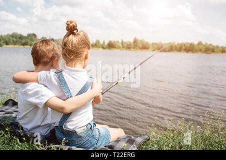 Wunderschöne Kinder sitzen in der Nähe von See und umarmten einander. Mädchen ist Angeln. Sie hält Fisch-rod. Sie alleine sind es Stockfoto