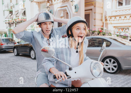 Aufgeregt Mädchen treibt motorcylcle. Sie ist sehr glücklich darüber. Kerl sitzt hinter ihr und schaut frustraed. Junger Mann, Hände auf Helm Stockfoto