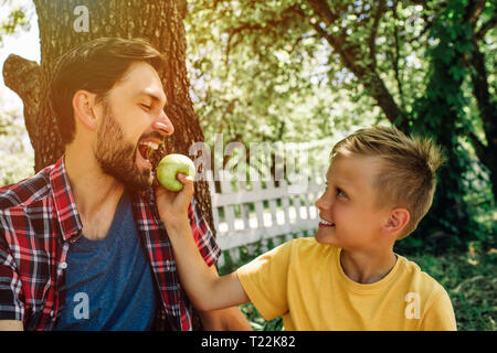 Nettes Bild von Vater und Sohn zusammen sitzen outsode unter dem Baum. Junge hält Apple während sein Vati Beißen ist Stück von es Stockfoto