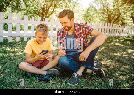 Kleiner Junge sitzt auf Gras mit seinem Vati mit gekreuzten Beinen. Kind hält das Telefon in der Hand und es zu betrachten. Er lächelt. Papa ist auf der Suche nach einem Stockfoto