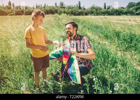 Kind steht, während sein Vater in squatt Position sitzt. Sie halten Kite. Jungs wollen, gemeinsam zu spielen und dafür zu sorgen, dass er in den Himmel fliegen Stockfoto
