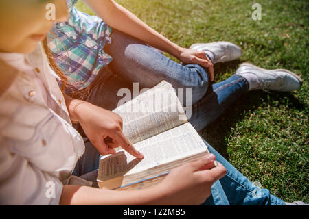 Schnittansicht der kleinen Mädchen sitzen mit Erwachsenen auf dem Gras. Es ist sonnig draussen. Mädchen ist ein Buch lesen und zeigen Sie auf Wörter. Nach hält die Hand auf Stockfoto