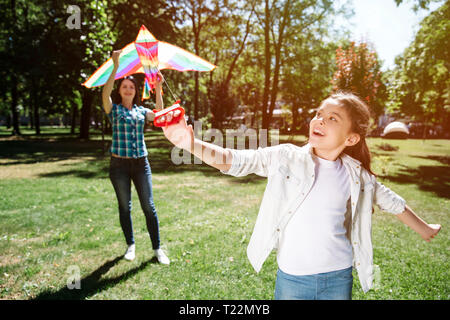 Ein weiteres Bild von Mädchen spielen mit Kite mit ihrer Mama. Mädchen steht, die sich in einem von vorn und ziehen Thread von Kite. Frau hält Kite und zu versuchen, Stockfoto