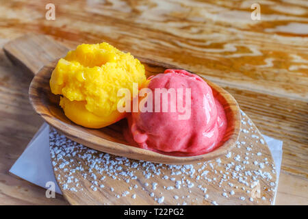 Mango und Himbeere Obst Sorbet Kugeln in eine hölzerne Schüssel auf einer Servierplatte mit Puderzucker auf einen Holztisch mit einer interessanten Holzmaserung surf sprengte Stockfoto
